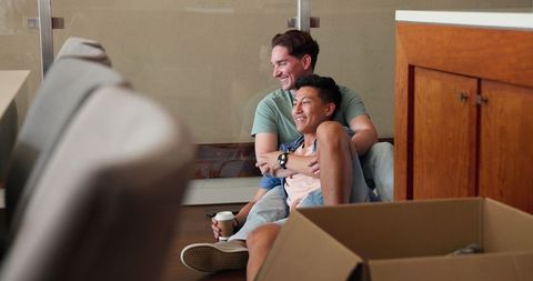 Happy Multicultural Gay Couple Relaxing Amid Moving Boxes at Home