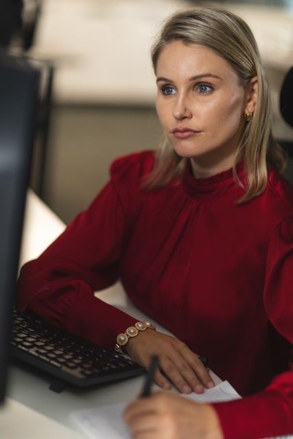 Caucasian businesswoman working late in the evening in a modern office, sitting at a desk, using a desktop computer, taking notes.
