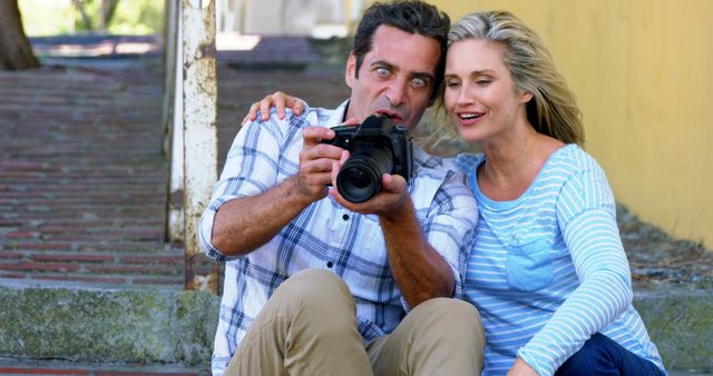 Smiling couple sitting on steps, enthusiastically reviewing photos on a DSLR camera. Ideal for use in promoting photography courses, outdoor activities, leisure time, emotional expressions, and happy moments shared between couples.