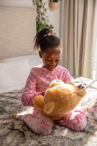 African american girl holding brown teddy bear while wearing pink zigzag pajamas on bed