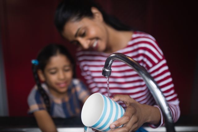 Smiling woman with daughter washing cup at kitchen sink at home