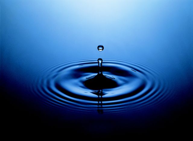 Depicts single water droplet creating ripples on a calm blue surface. Can be used for articles and advertisements related to tranquility, meditation, environmental conservation, purity, or fluid dynamics.