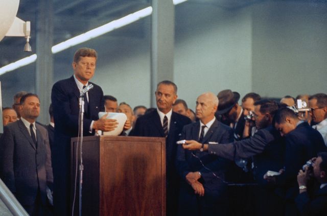 President John F. Kennedy is delivering a speech at a NASA site in 1962. Vice President Lyndon B. Johnson, NASA Administrator James E. Webb, and Dr. Robert R. Gilruth are also present. The audience consists of media and employees. This image showcases significant moments in space exploration history and leadership, making it perfect for historical documentation, presentations on space programs, and educational content about U.S. history and political figures.