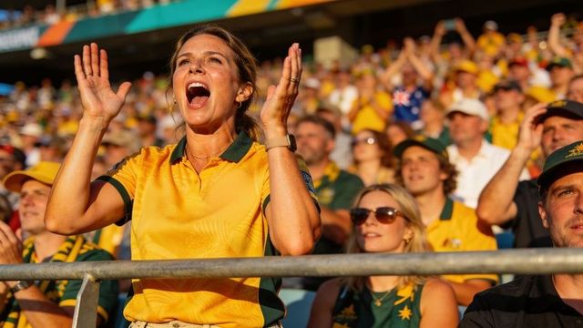 Cheering woman in yellow jersey raising hands front-row stadium crowd in sunlight
