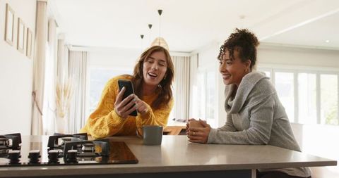 Diverse female friends sharing smartphone and coffee at modern sunlit kitchen island, chat