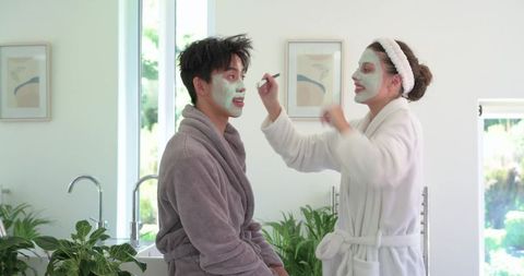 Diverse couple applying green facial masks while laughing in bright minimalist bathroom