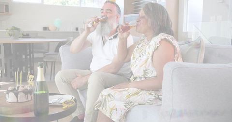 Sunlit senior couple toasting champagne on sofa with birthday cake in modern living room