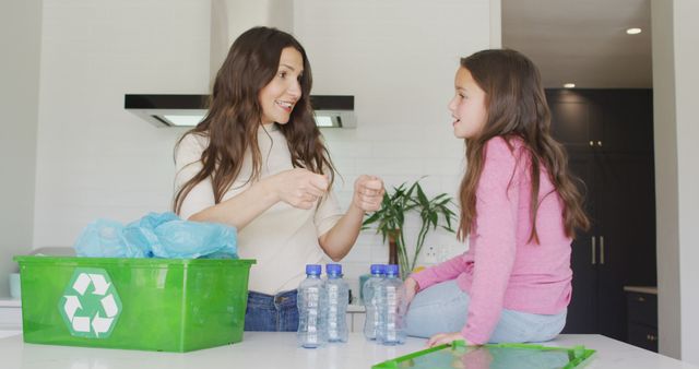 Mother and daughter are discussing recycling in a modern kitchen with a green recycling bin filled with plastic bottles on the counter. This image can be used for themes related to family bonding, environmental awareness, eco-friendly practices, and home recycling activities.