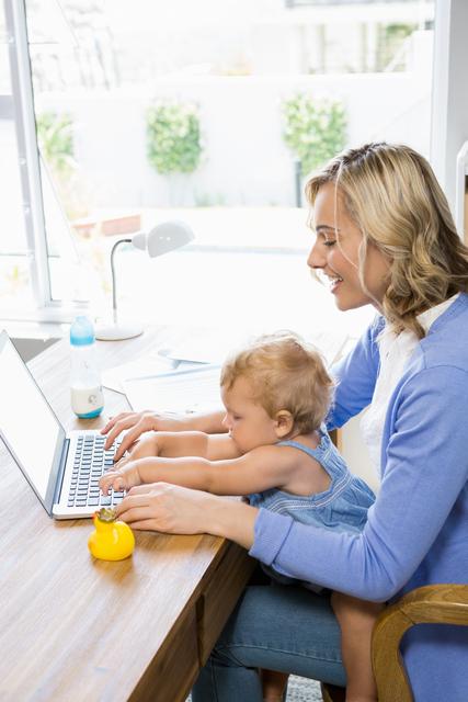Mother and baby girl sitting at a wooden table, both using a laptop. The mother is smiling while the baby is also engaged with the laptop. A baby bottle and a rubber duck are on the table. Bright and airy home environment with natural light coming through the window. Ideal for use in articles about work-life balance, parenting, remote work, and family technology use.