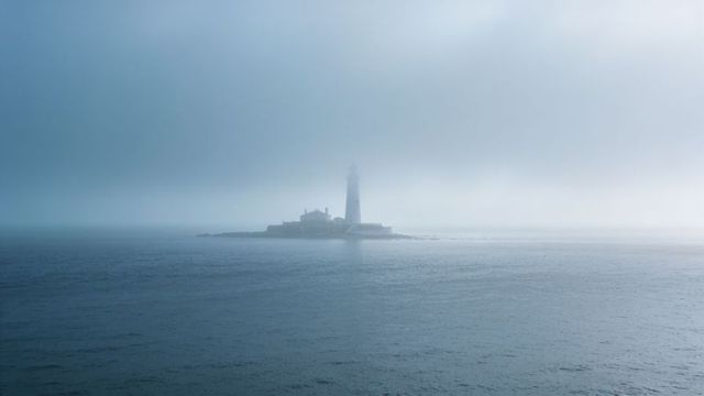Misty lighthouse rising from rocky islet, guiding ships through foggy sea horizon