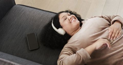 Smiling woman relaxing on sofa listening to music with headphones and smartphone