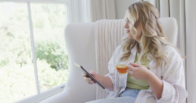 Young woman sitting comfortably by a window, holding a tablet in one hand and sipping tea with the other. She appears relaxed and leisurely, surrounded by natural light. This image is ideal for concepts related to relaxation, technology use at home, and lifestyle.