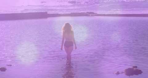 Backlit woman walking into violet sea at sunset, wading toward distant horizon