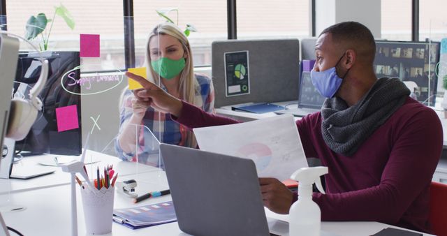 Two business colleagues engaging in a discussion in a modern office. Both are wearing face masks and there is a sanitizer spray bottle on the desk, indicating they are following health and safety protocol. Collaborating and pointing at documents and sticky notes on clear dividers. Suitable for topics such as workplace safety, collaboration during the pandemic, and modern office settings.