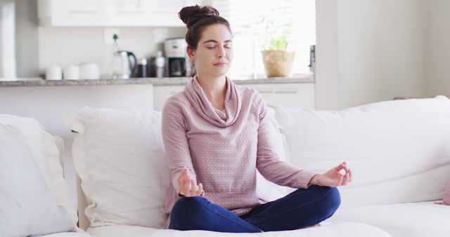 Young woman meditating in lotus position on white couch in bright living room. Peaceful environment enhances the mood of calm and serenity. Suitable for blogs and articles on mindfulness practices, mental health, and indoor wellness routines.
