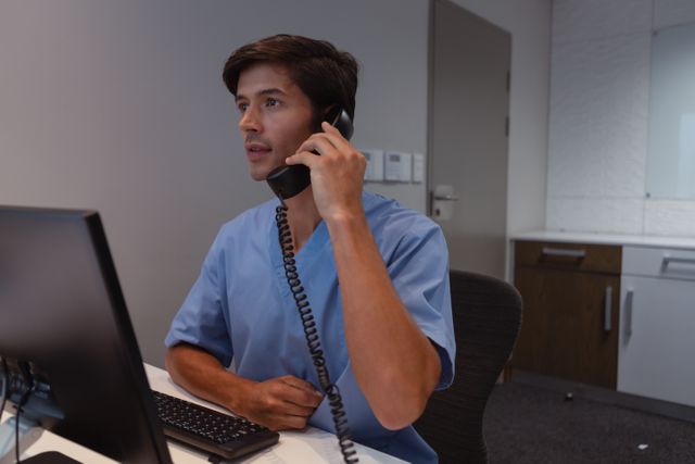Male surgeon in blue scrubs talking on a landline phone while sitting at a desk with a computer in a hospital office. Ideal for use in healthcare, medical, and professional communication contexts. Can be used in articles, websites, and promotional materials related to healthcare services, hospital administration, and medical professionals.