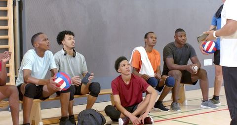 Diverse high school volleyball players listening to coach on gym bench preparing for practice