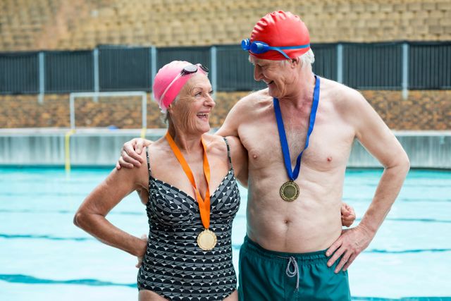 Senior couple standing by the poolside, proudly wearing medals around their necks. They are smiling and appear happy, showcasing their achievements in swimming. This image can be used to promote active lifestyles for seniors, health and fitness programs, or sports events for the elderly.