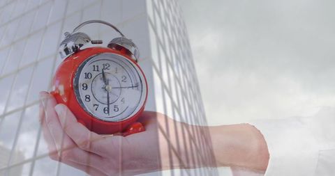 Holding red alarm clock over glass office tower representing time management and urgency