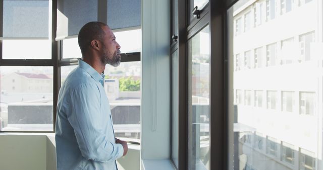 Middle-aged man standing in an office, looking pensively out the window at urban buildings. Ideal for concepts of reflection, contemplation, inspiration, work-life balance, and modern urban environments.