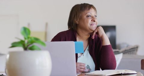 Midlife woman gazing thoughtfully while working from home with laptop and notebook