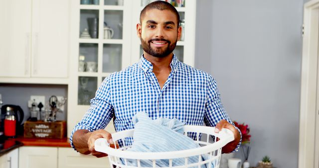 A young Asian man smiles while holding a laundry basket filled with clothes, with copy space. His casual attire and the home setting suggest a comfortable, domestic routine.