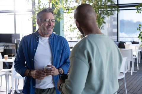 Casual coworkers chatting with coffee in sunlit modern office lounge during break