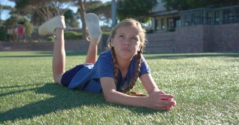 Preteen girl lying on green turf holding small red toy looking thoughtful at school field