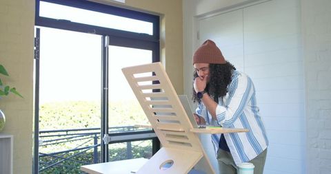 Young man leaning over laptop at standing desk in bright home office with balcony view