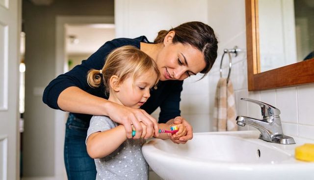 Mother guiding toddler brushing teeth at bathroom sink, daily parenting routine and bonding