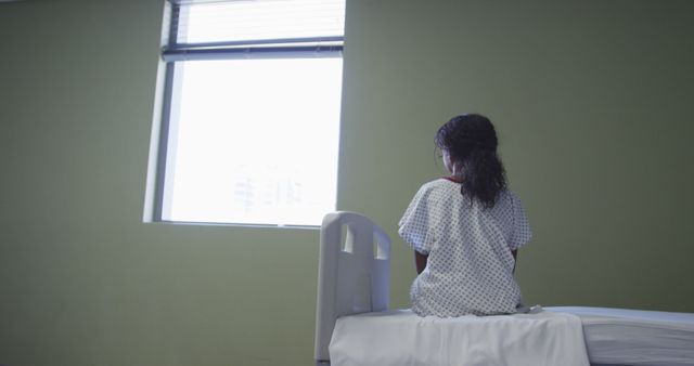 Biracial girl sitting up in hospital bed looking through the window. medicine, health and healthcare services.