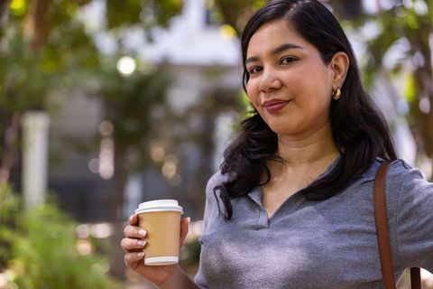 Confident woman enjoying takeaway coffee outdoors in urban green courtyard, smiling