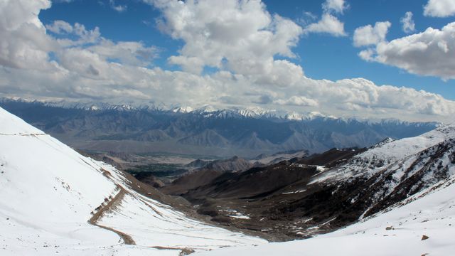 Breathtaking snow-covered mountain range under a vibrant blue sky filled with scattered clouds. A winding road can be seen cutting through the snow, leading to a valley below. Ideal for travel blogs, winter sports promotions, nature magazines, or websites focusing on outdoor adventures and pristine environments.
