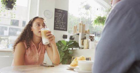 Young woman sipping iced latte at sunlit cafe window with croissant and greenery