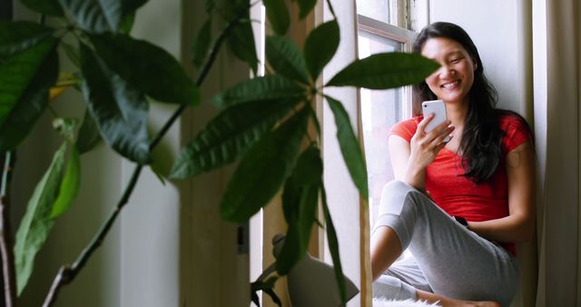 Young Woman Smiling While Using Smartphone by Window Plant Indoor Setting - Download Free Stock Images Pikwizard.com