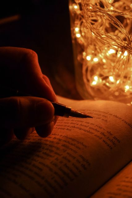 A hand holding a pen interacts with an open book, lit warmly by nearby fairy lights, creating a cozy and intimate atmosphere. This image is ideal for use in articles or advertisements focused on reading, relaxation, nocturnal routines, literary environments, or cozy indoor settings.