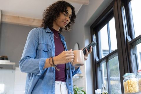 Non-binary adult standing in sunlit modern kitchen holding mug and checking phone by countertop jars