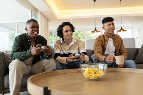 Friends playing video games on couch holding controllers and enjoying snacks in bright living room