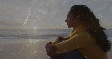 Contemplative woman sitting on beach hugging knees, gazing at sunset with film texture