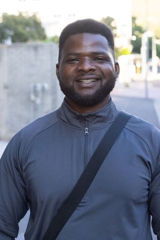 Smiling african man standing on city sidewalk wearing dark-gray pullover and crossbody strap