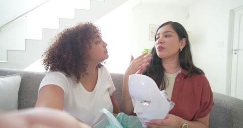 Diverse female friends sharing led facial mask on grey sofa at home