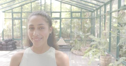 Smiling woman tending potted plants in sunlit glass greenhouse for urban gardening at home
