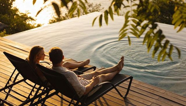 Couple relaxing on lounge chairs by infinity pool at sunset, barefoot on deck, sunlit