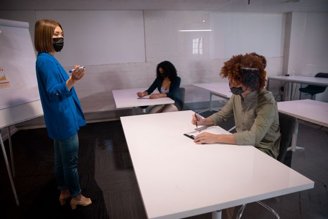 Businesswoman presenting to colleagues in an office setting, all wearing face masks for safety during the COVID-19 pandemic. Ideal for illustrating modern workplace practices, health and safety measures, and professional teamwork during a pandemic. Useful for articles on business continuity, workplace safety, and adapting to new norms.