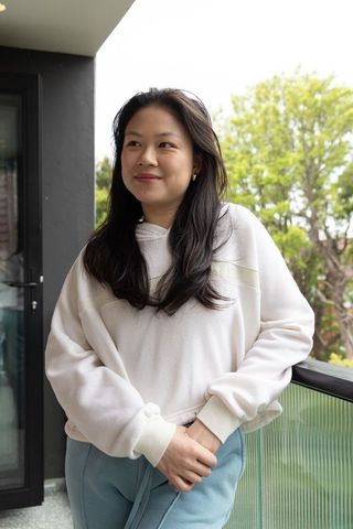 Asian woman standing on balcony wearing white hoodie and blue pants, clasping hands, serene lifestyl
