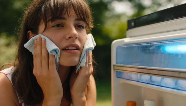 Young woman cooling face with chilled cloths by open refrigerator outdoors on hot day