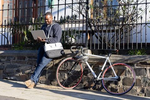 African american commuter leaning on wall working on laptop next to bicycle with satchel