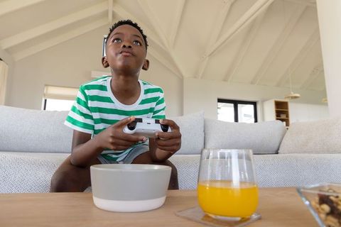 Young african american boy playing video game on couch with juice and snacks at home