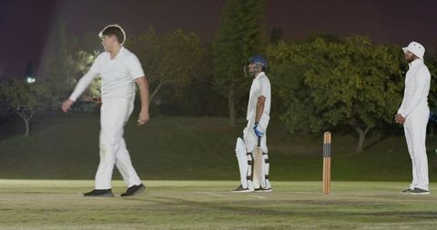 Night cricket match players standing and walking on illuminated pitch near stumps