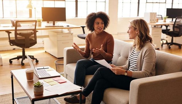 Two coworkers discussing on sofa in modern open office with laptop and papers